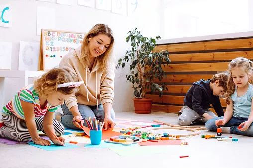 Porträt einer jungen lächelnden Lehrerin, die mit Kindern sitzt und ein Gemälde auf rotem Papier betrachtet.