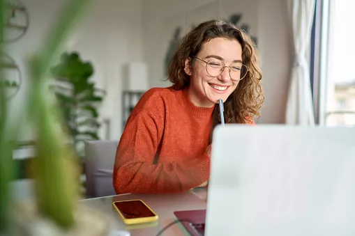 Junge glückliche Studentin benutzt Laptop, schaut sich ein Webinar an und schreibt zu Hause.